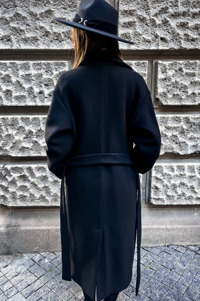 Devojka nosi crni kaput na vezivanje. Slikana je ispred sivog zida i drži ruke u džepovima.
