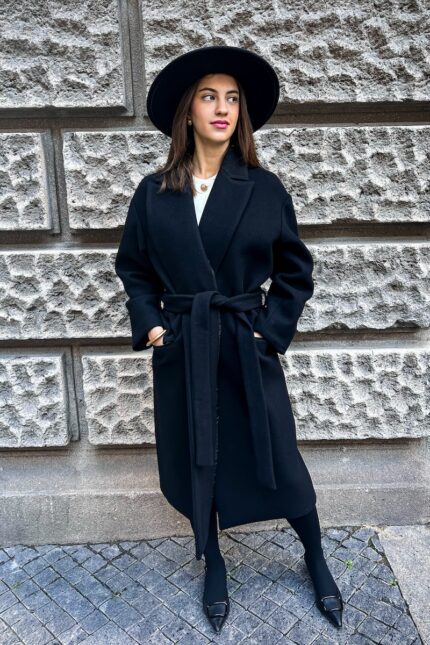 The girl is wearing a black tie-up coat with a black hat and black shoes. She is pictured in front of a gray wall.