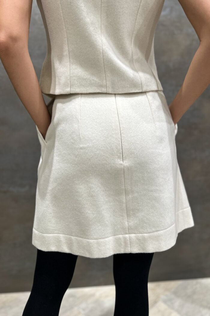 Kratka EMA suknja od krem bele vune sa faltama na prednjoj strani i ukrojenim džepovima, slikana otpozadi.