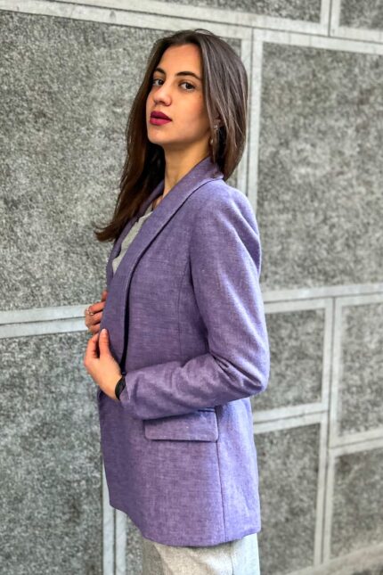 The girl is wearing a purple wool LETITIA blazer with gray pants and is standing in front of a gray wall.
