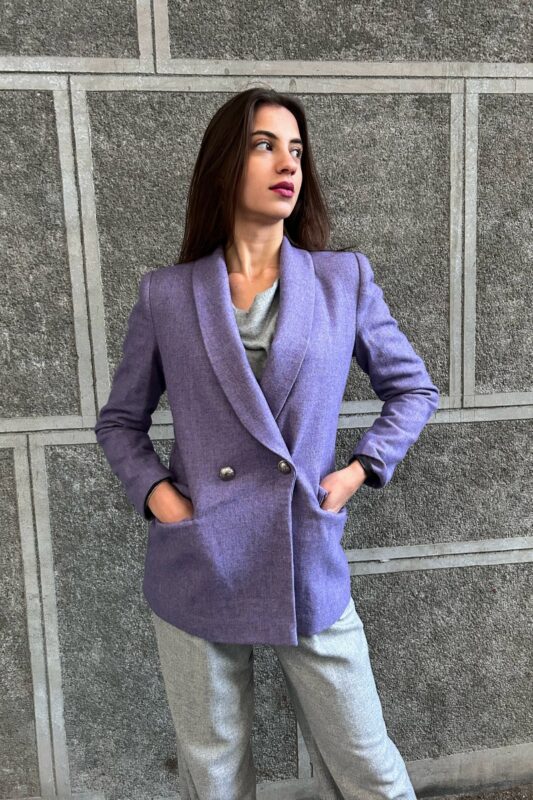 The girl is wearing a purple wool LETITIA blazer with gray pants and is standing in front of a gray wall..
