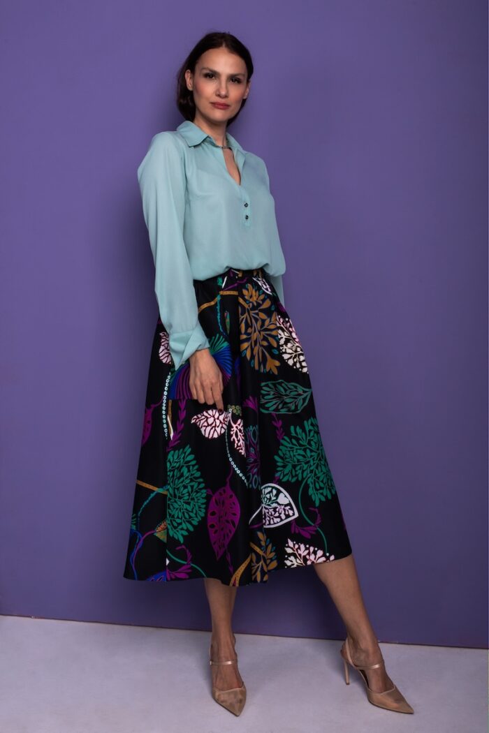 A woman stands in front of a purple background and wears a mint green blouse with a black patterned skirt.