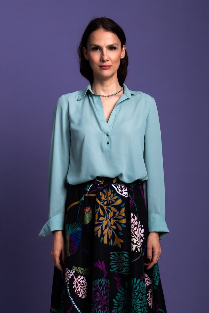 A woman stands in front of a purple background and wears a mint green blouse with a black patterned skirt.