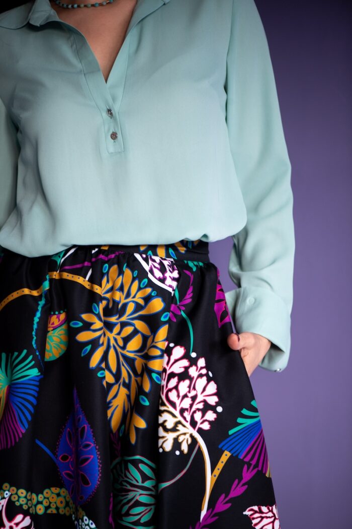 A woman stands in front of a purple background and wears a mint green blouse with a black patterned skirt