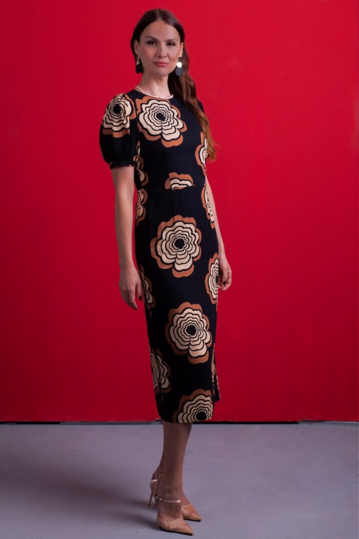 A woman wears a black dress with floral motifs, standing in front of a red background.