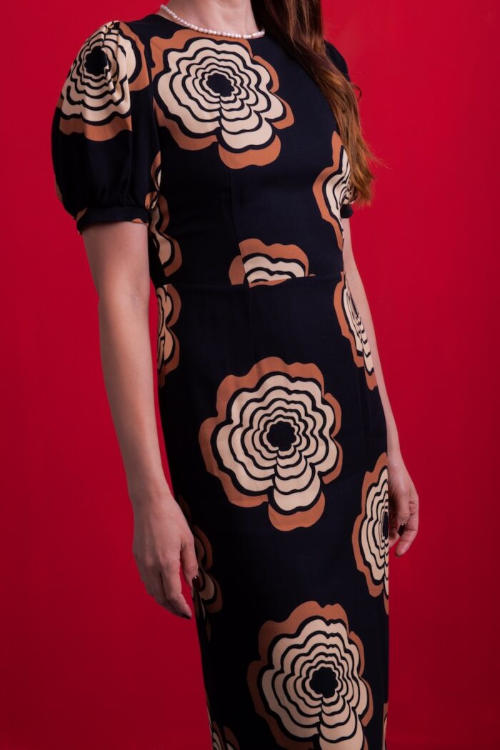 A woman wears a black dress with floral motifs, standing in front of a red background.