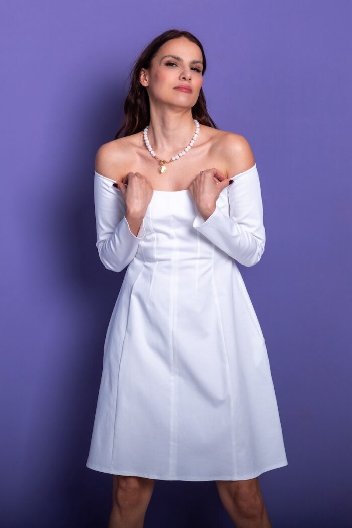 Žena nosi kratku belu haljinu i stoji ispred ljubičastog zida.