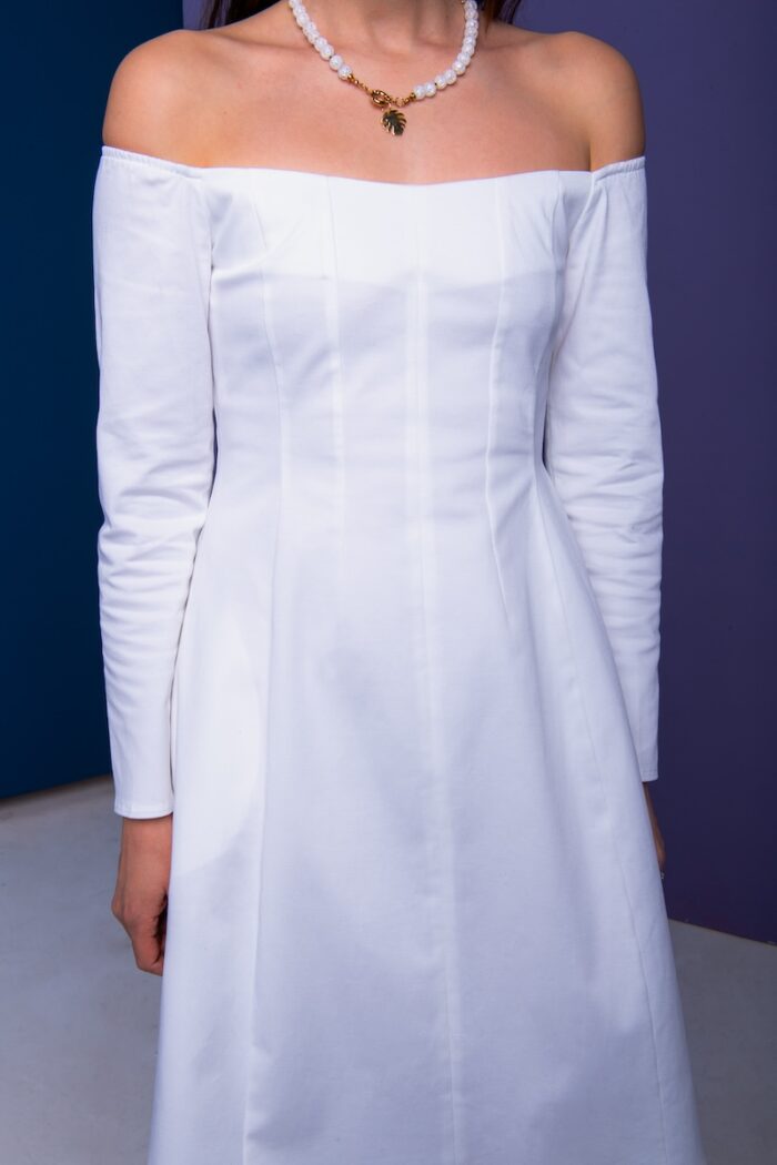 Žena nosi kratku belu haljinu i stoji ispred ljubičastog zida.