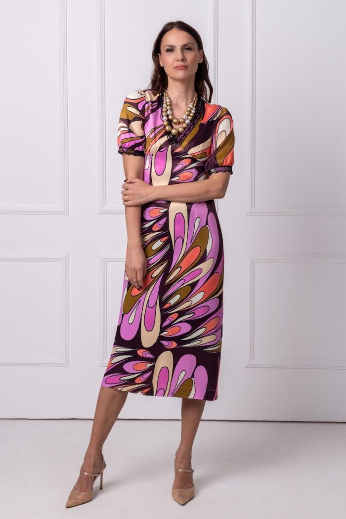 The woman wears a colorful midi dress with short sleeves and a V-neckline. She is standing in front of a white wall.