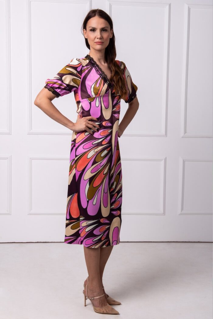 The woman wears a colorful midi dress with short sleeves and a V-neckline. She is standing in front of a white wall.