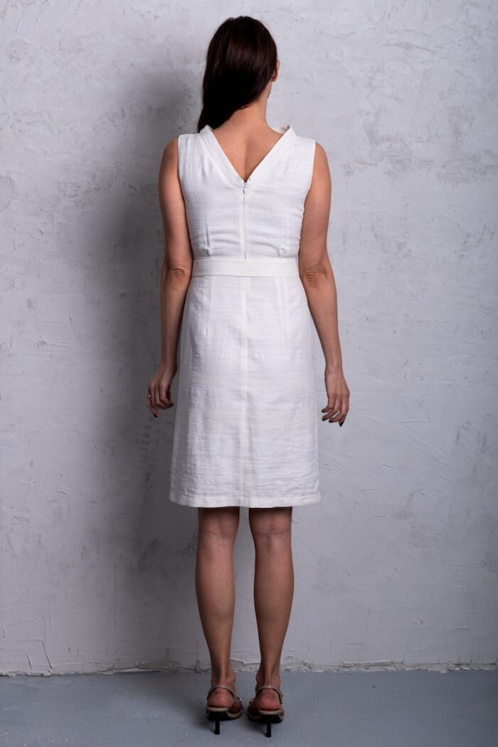 A woman wears a white sleeveless dress made of shantung silk and stands in front of a gray wall.