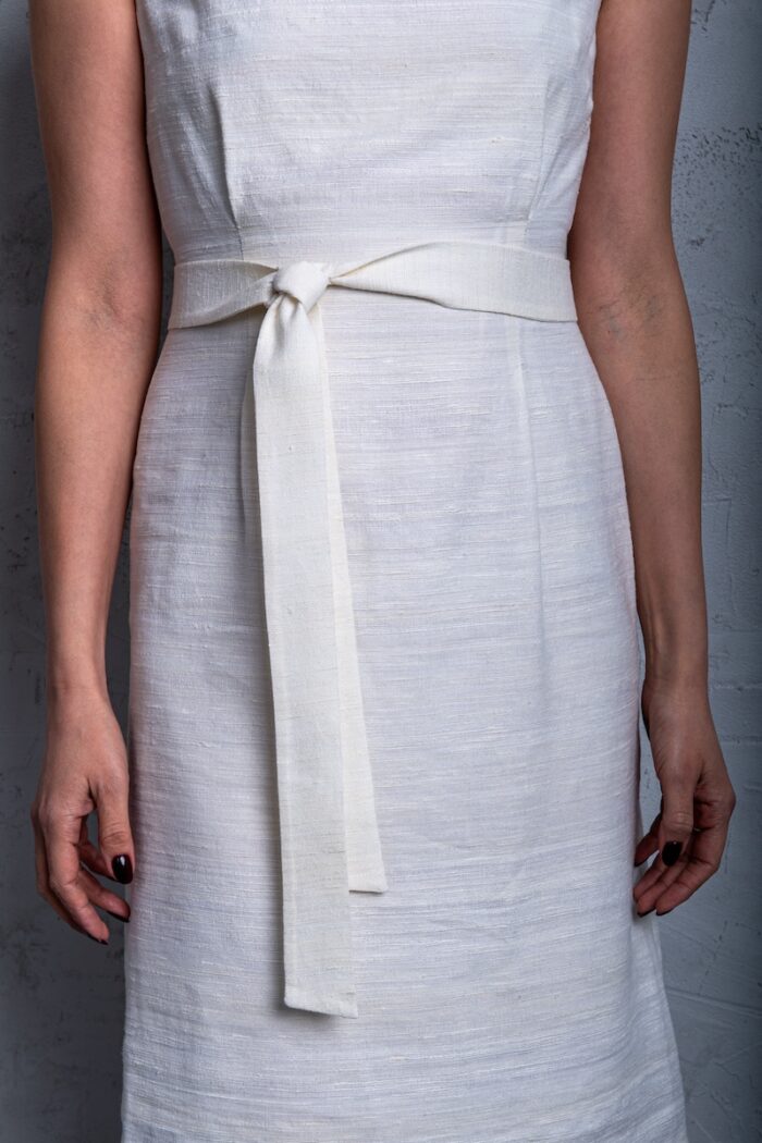Žena nosi belu haljinu bez rukava od šantung svile i stoji ispred sivog zida.