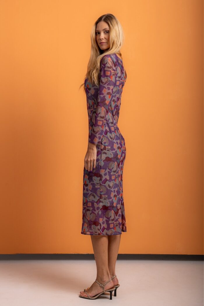 Plava devojka nosi dezeniranu haljinu midi dužine i stoji ispred narandžaste pozadine.
