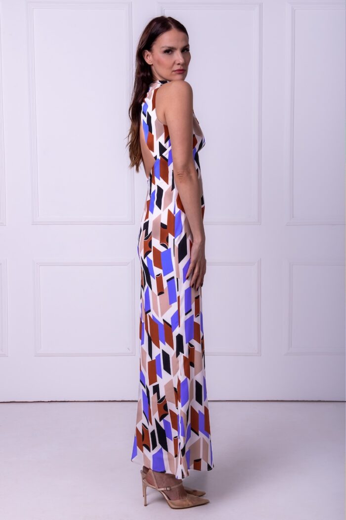 A woman wears a long silk patterned sleeveless dress and stands in front of a white wall.