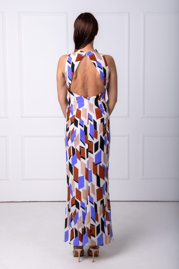 A woman wears a long silk patterned sleeveless dress and stands in front of a white wall.