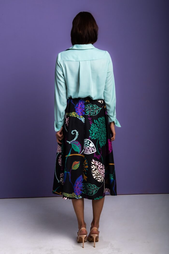 The woman is wearing a black skirt with a floral pattern, along with a green shirt. She stands in front of a purple background.