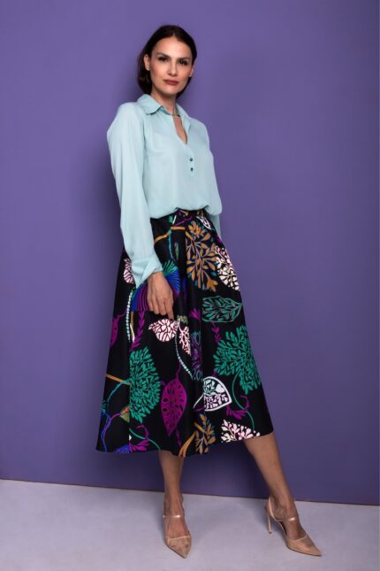 The woman is wearing a black skirt with a floral pattern, along with a green shirt. She stands in front of a purple background