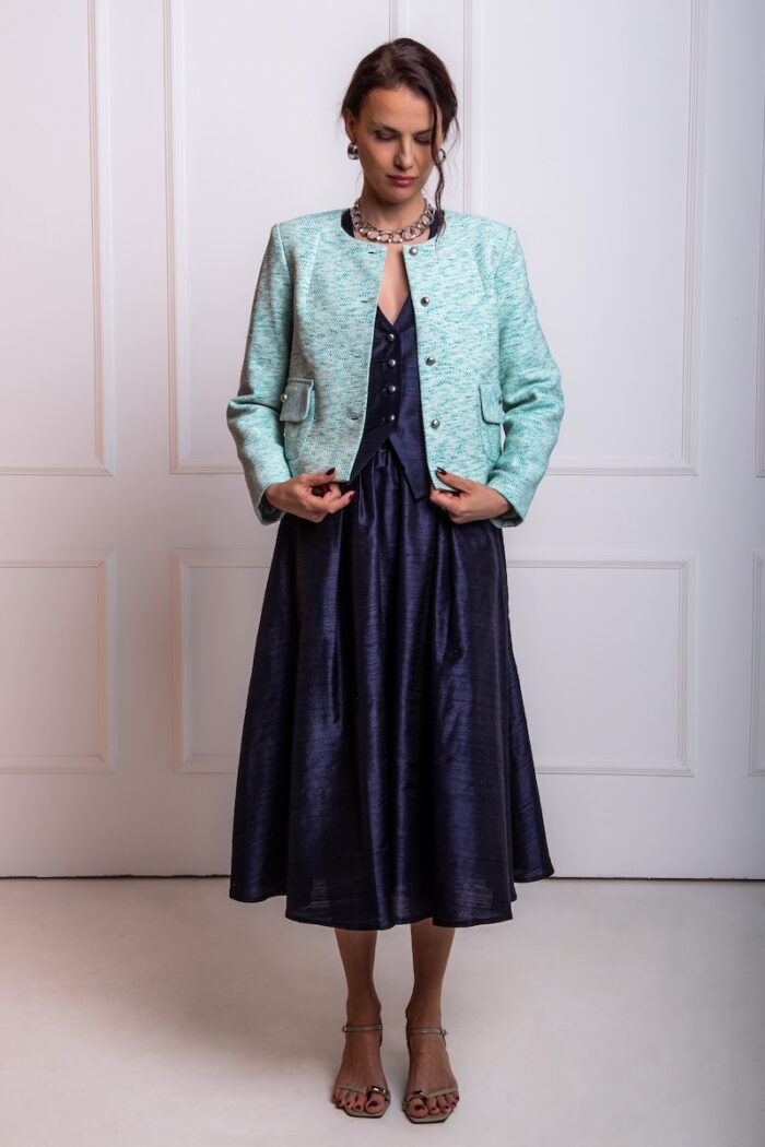 The woman wears a navy shantung silk midi skirt paired with a turquoise boucle blazer. She is standing in front of a white wall.