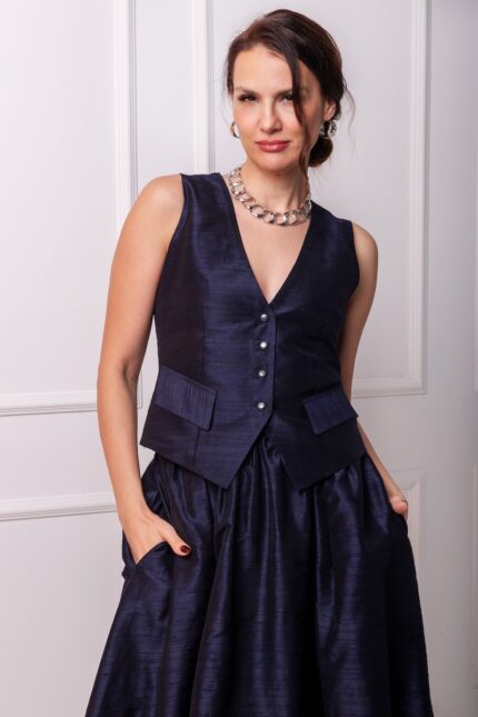 The woman wears a navy vest and a navy skirt of the same material. She is standing in front of a white wall.