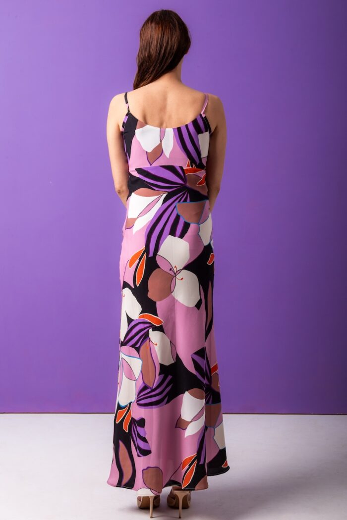 The woman wears a long, silk dress with a floral pattern. He is standing in front of a purple background.