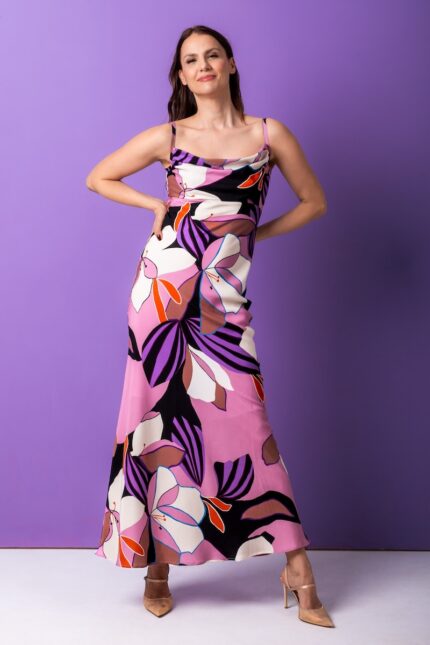 Žena nosi dugu, svilenu haljinu floralnog dezena. Stoji ispred ljubičaste pozadine.