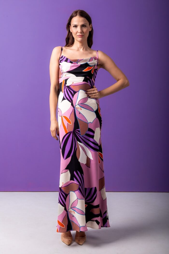 The woman wears a long, silk dress with a floral pattern. He is standing in front of a purple background.