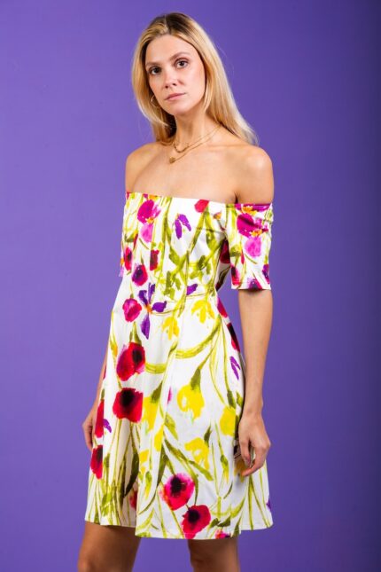 A blonde girl wears a short floral dress and stands in front of a purple background.