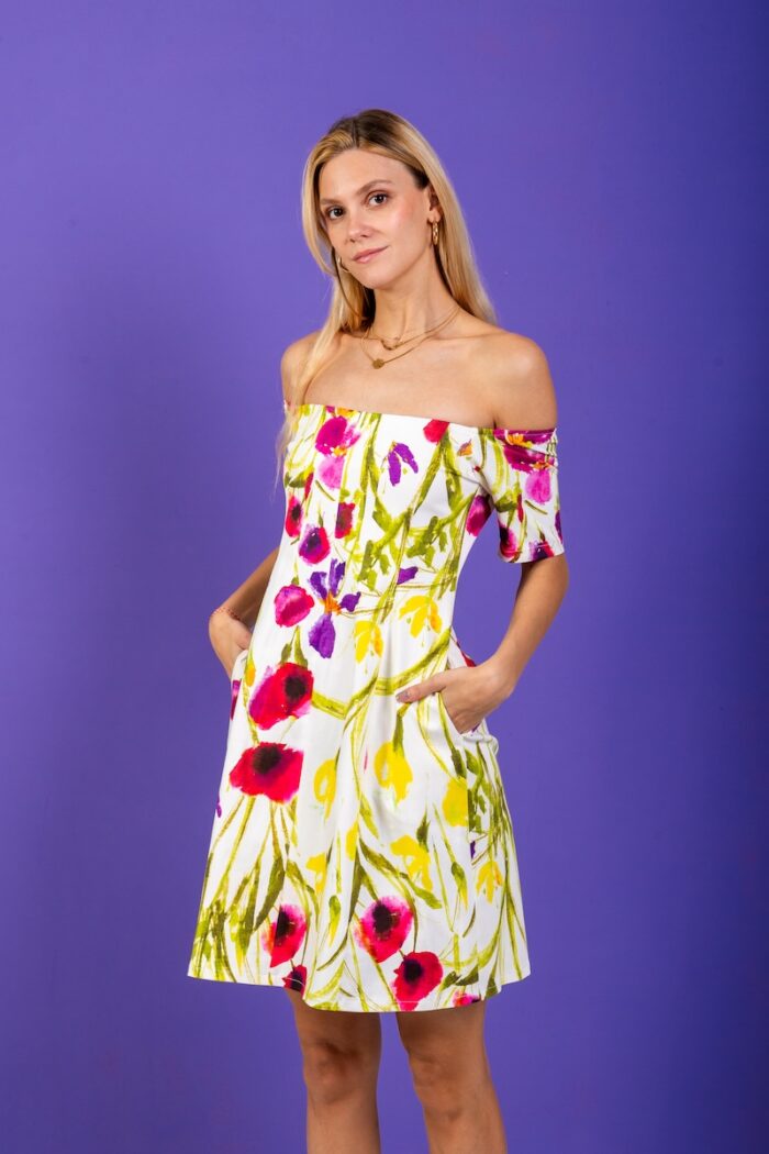 A blonde girl wears a short floral dress and stands in front of a purple background.