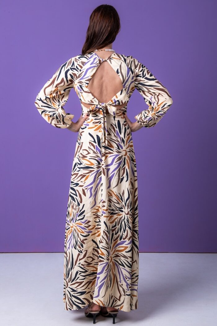 The woman wears a long patterned dress with long sleeves. He is standing in front of a purple background.