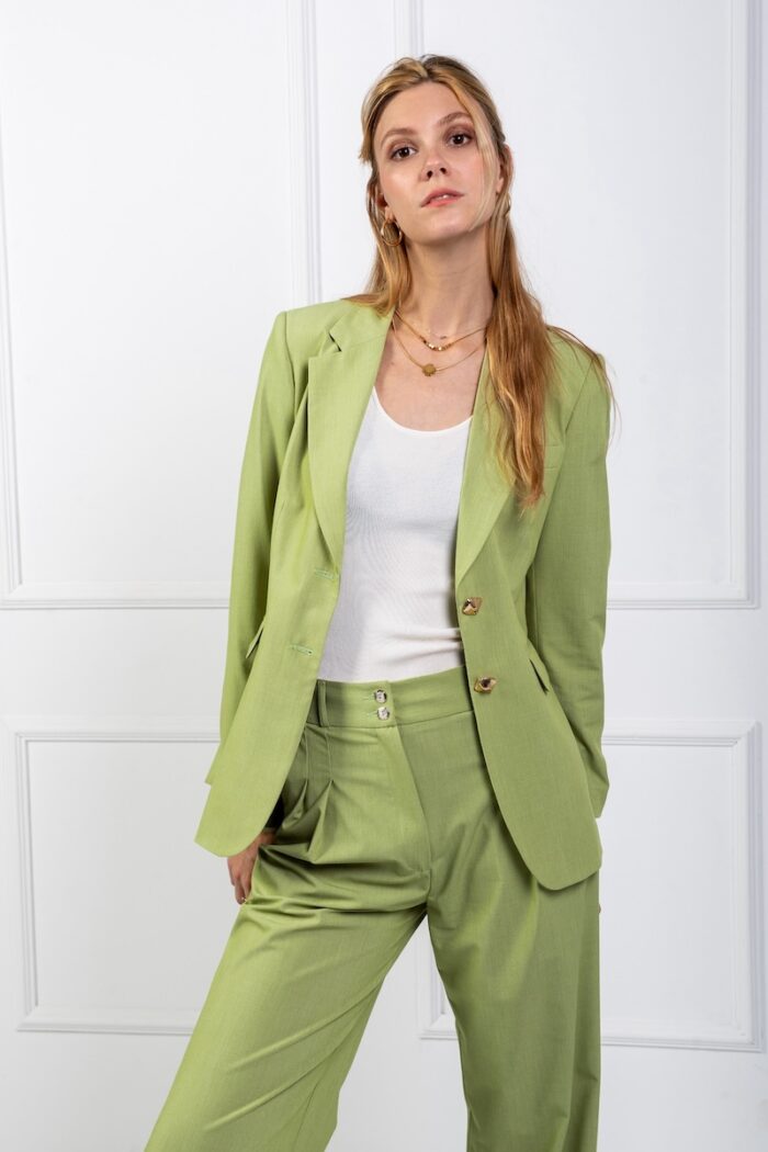 A blonde girl wears a green blazer and stands in front of a white wall.