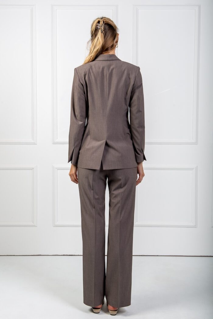 The blonde girl wears a waisted brown blazer and skinny pants of the same material. She is standing in front of a white wall.