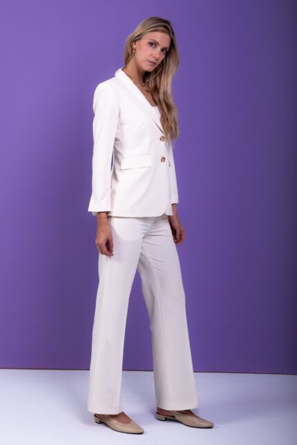 A blonde girl wears a white blazer and pants set and stands in front of a purple background.