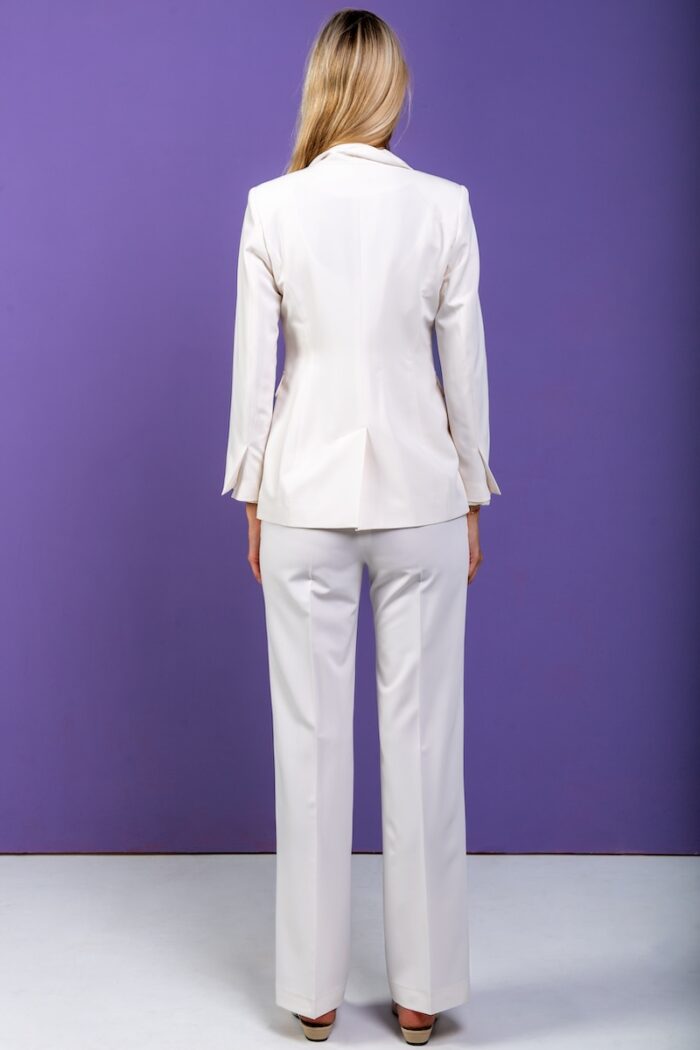 A blonde girl wears a white blazer and pants set and stands in front of a purple background.