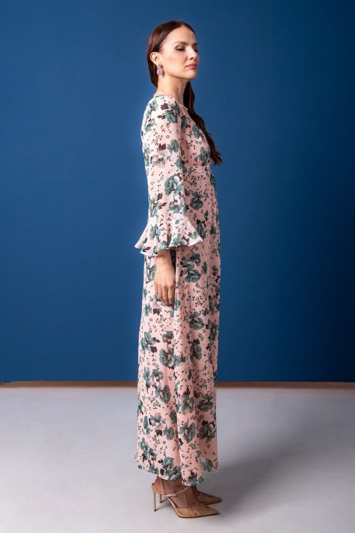 Žena nosi dugu haljinu cvetnog dezena i stoji ispred plave pozadine.