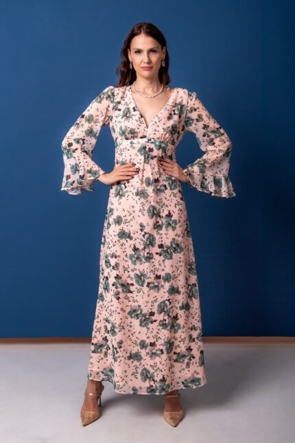 A woman wears a long floral patterned dress and stands in front of a blue background.