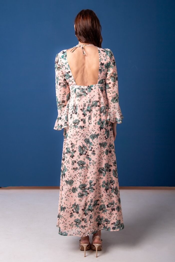 Žena nosi dugu haljinu cvetnog dezena i stoji ispred plave pozadine.