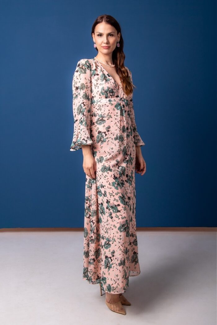 A woman wears a long floral patterned dress and stands in front of a blue background.