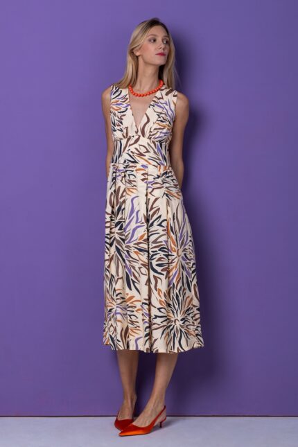 A blonde girl wears a sleeveless midi patterned dress and stands in front of a purple background.