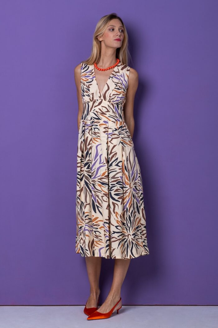 A blonde girl wears a sleeveless midi patterned dress and stands in front of a purple background.