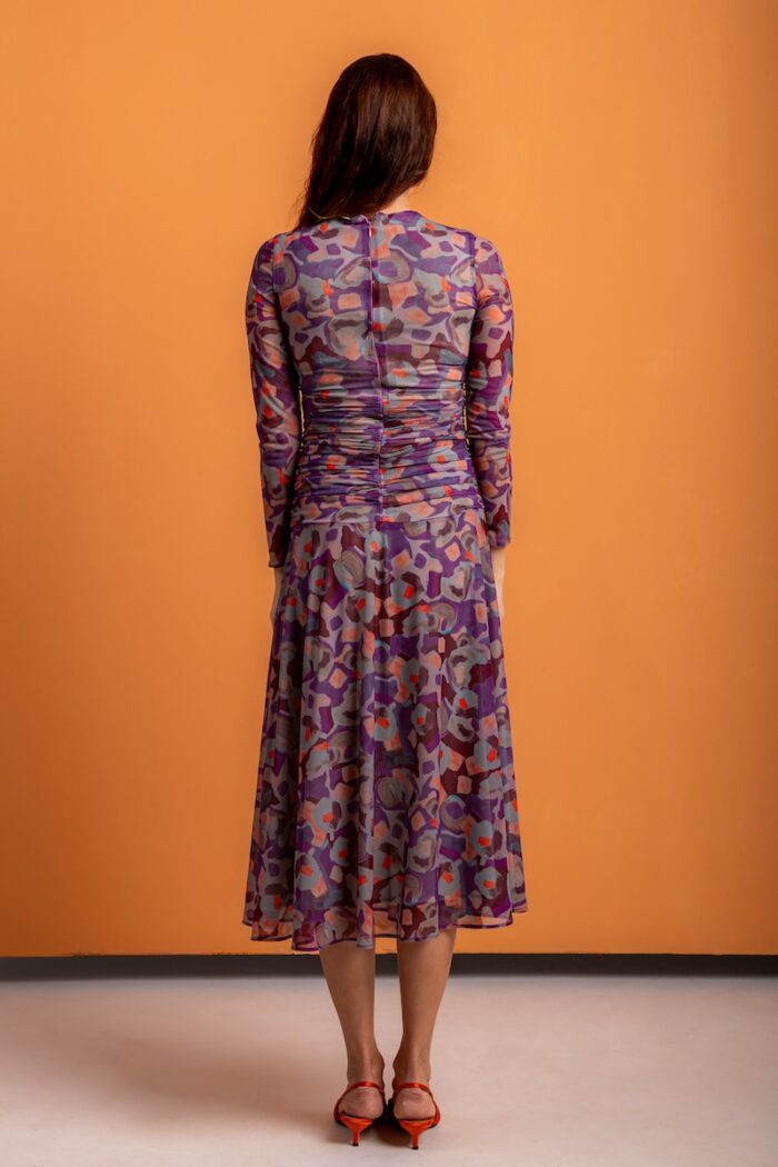 The woman wears a patterned midi dress with long sleeves and stands out against an orange background.