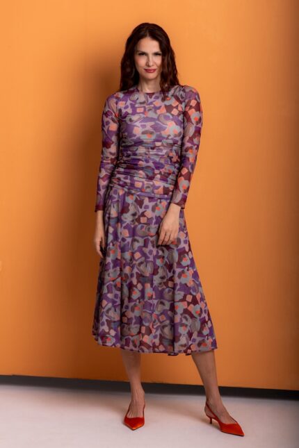 The woman wears a patterned midi dress with long sleeves and stands out against an orange background.
