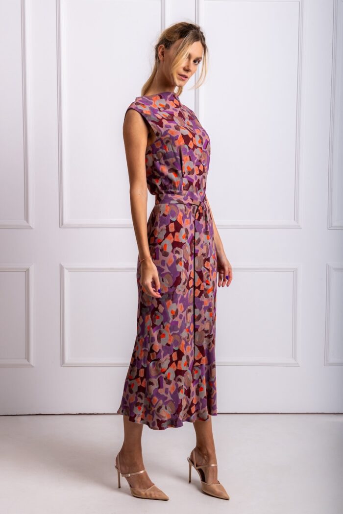 The blonde girl wears a patterned purple sleeveless midi dress. She is standing in front of a white wall.