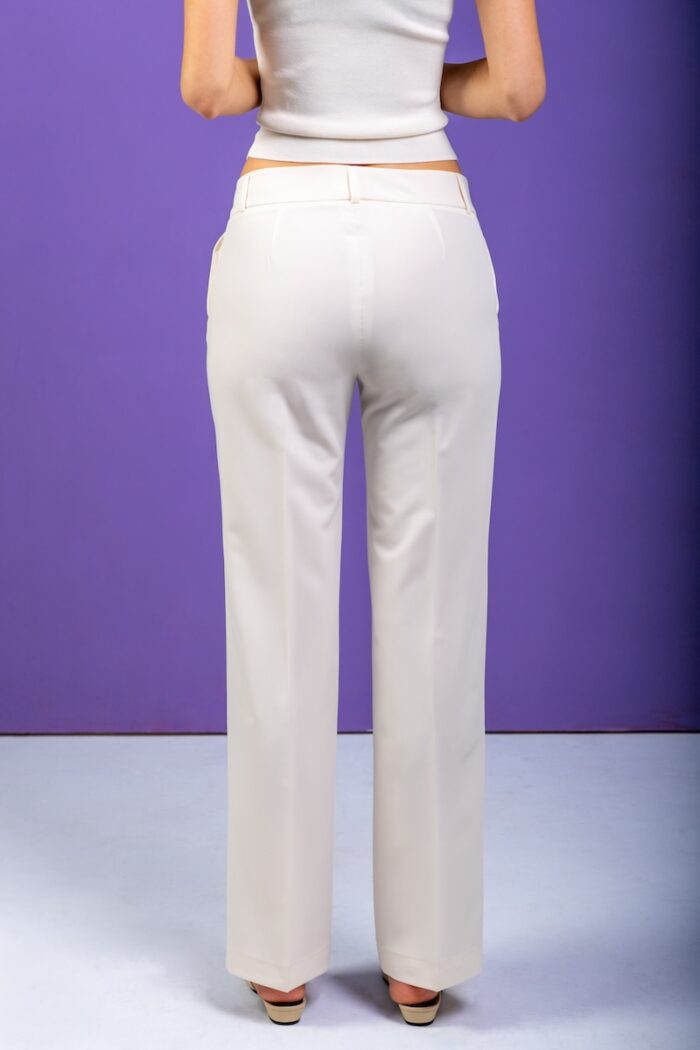 The girl wears white slim pants and a white blazer and stands in front of a purple background.
