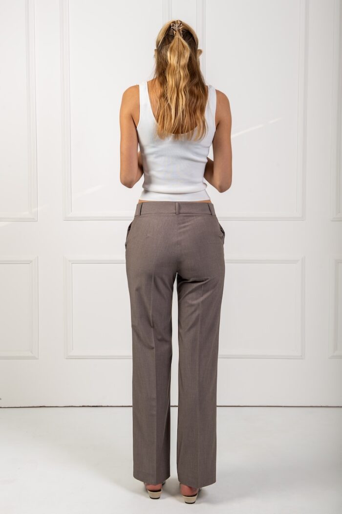 The blonde girl wears tight brown pants and a white strappy top. She is standing in front of a white wall.