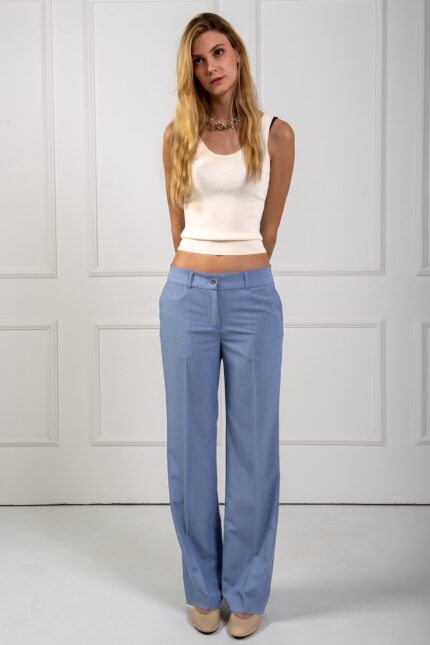 The blonde girl is wearing slim blue pants and a white top. She is standing in front of a white wall.