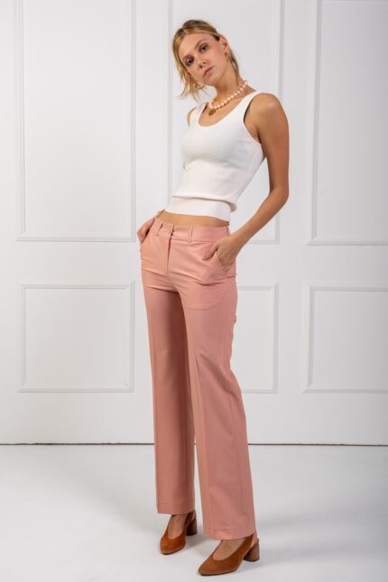 The blonde girl wears tight peach pants and a white strappy top. She is standing in front of a white wall.