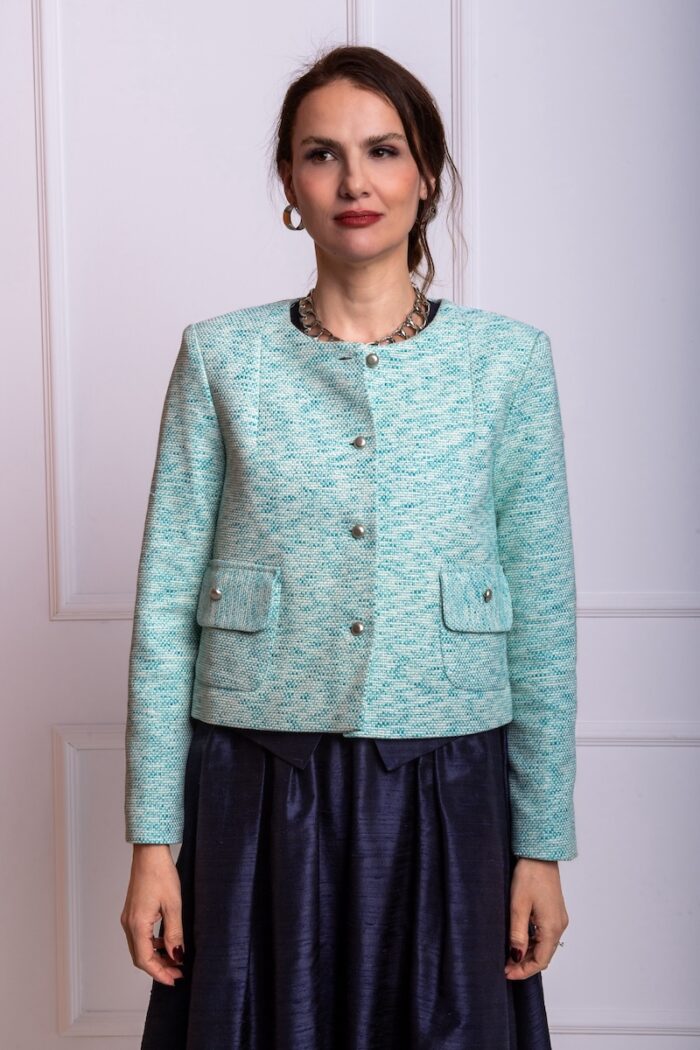 The woman wears a short, turquoise cotton bouclé blazer. She is standing in front of a white wall.