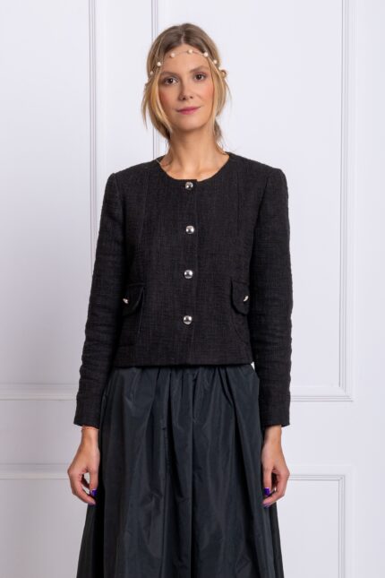 The blonde girl wears a black cotton blazer and a black midi skirt. She is standing in front of a white wall.