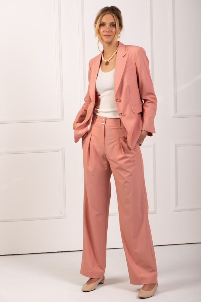 The blonde girl wears peach pants with wide legs and a white top. She is standing in front of a white wall.