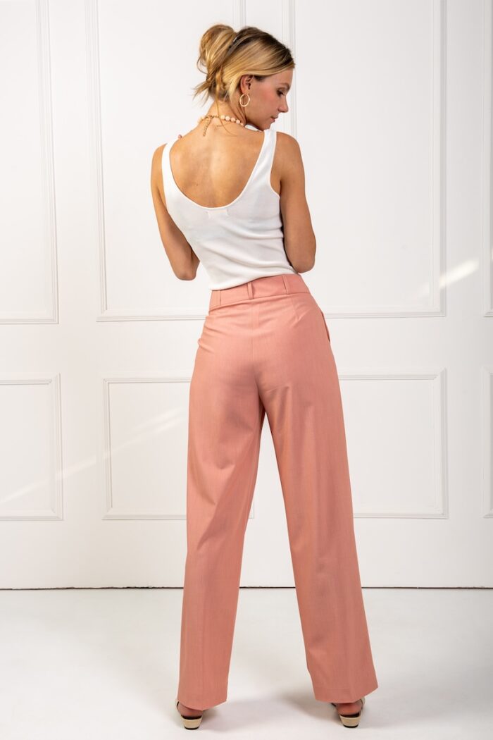 The blonde girl wears peach pants with wide legs and a white top. She is standing in front of a white wall.